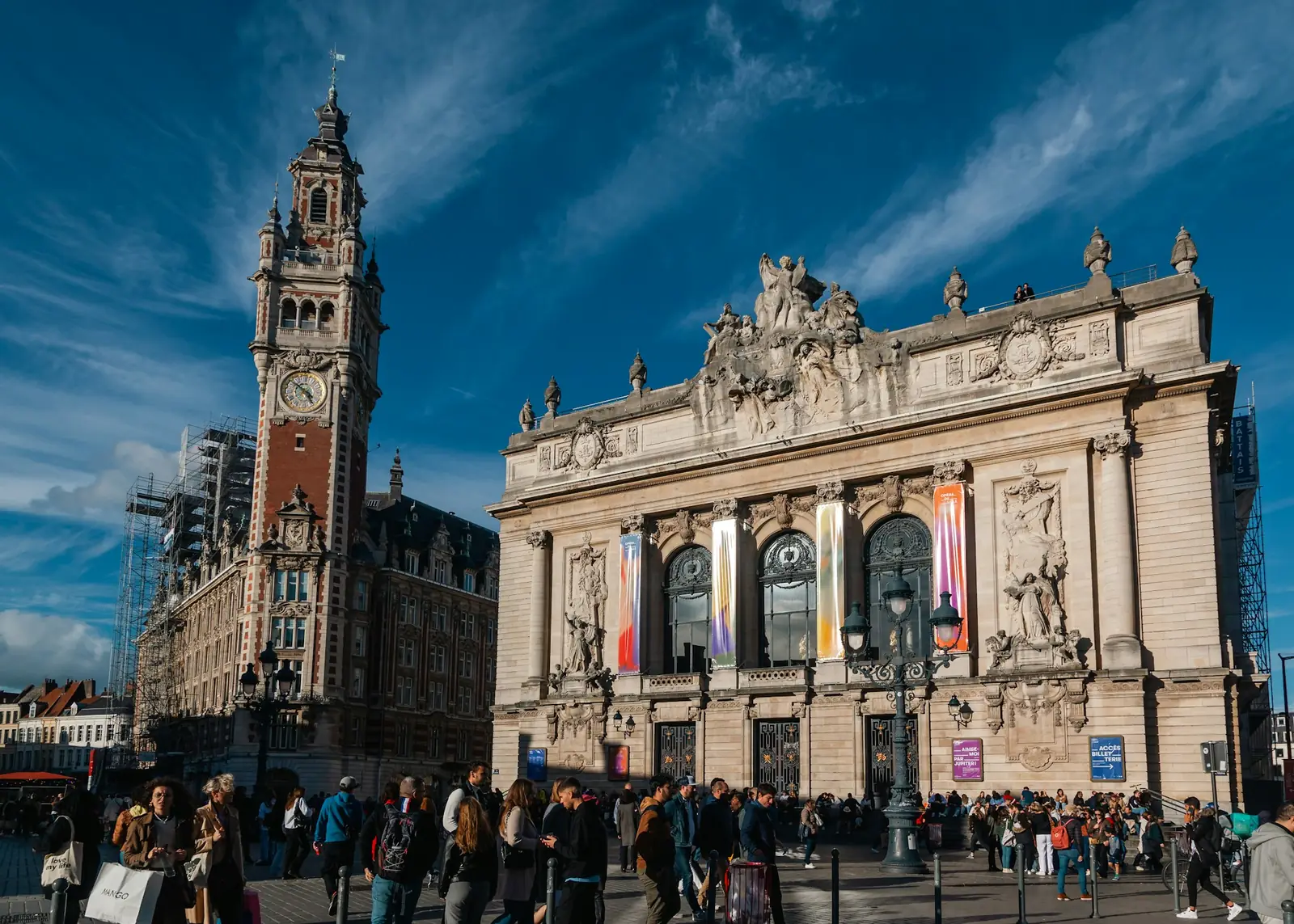 Des passants sur une place ensoleillée de Lille, devant un bâtiment à tour d'horloge